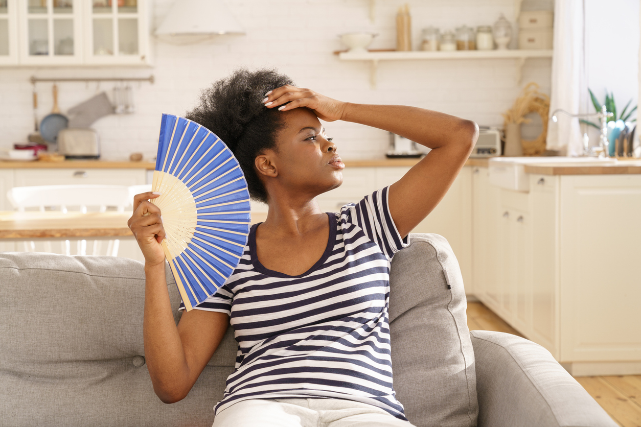 A woman sitting on a couch fans herself while holding her forehead, appearing overheated—illustrating discomfort caused by uneven home temperatures and poor indoor climate control.